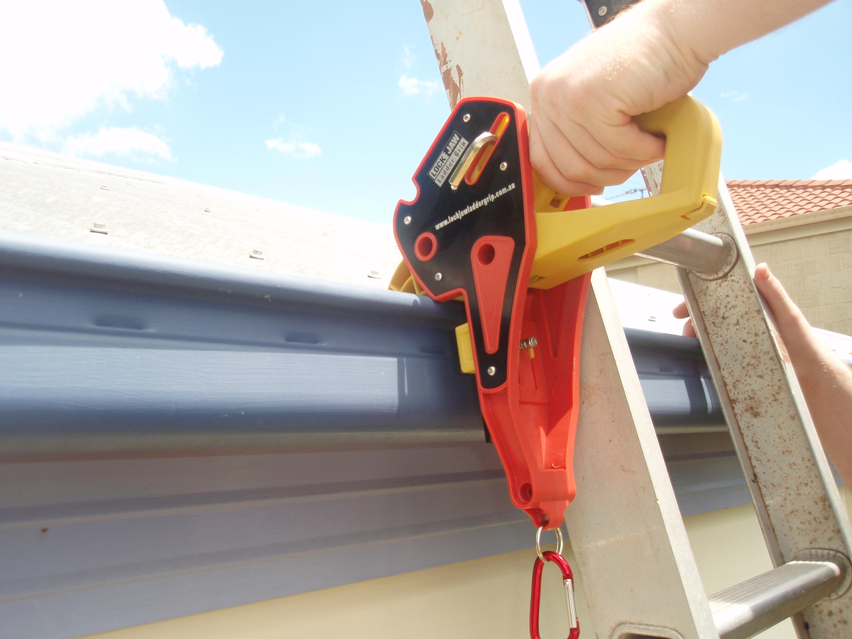 A hand secures a Lock Jaw Ladder Grip onto a ladder against a gutter.