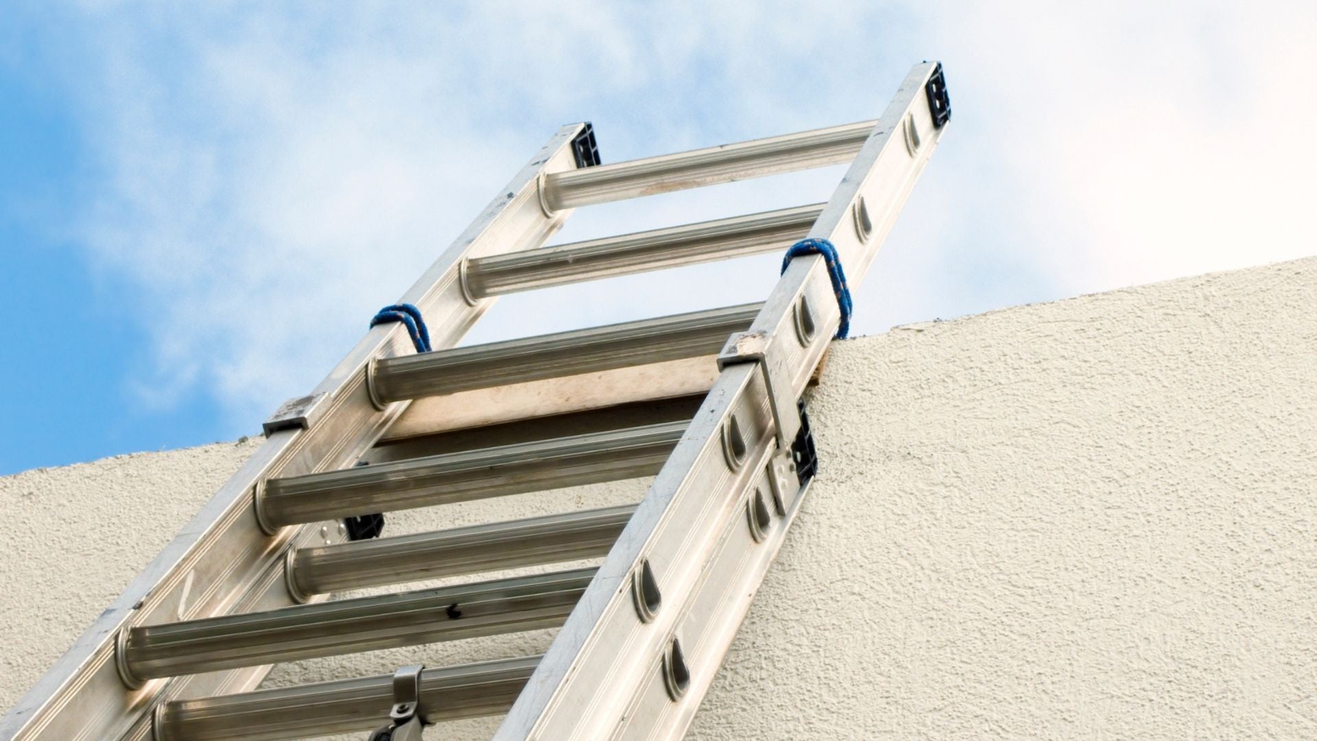 A triple ladder, possibly a Werner 24ft Type I Aluminum D-rung Extension Ladder, is shown leaning against a white wall under a blue sky.
