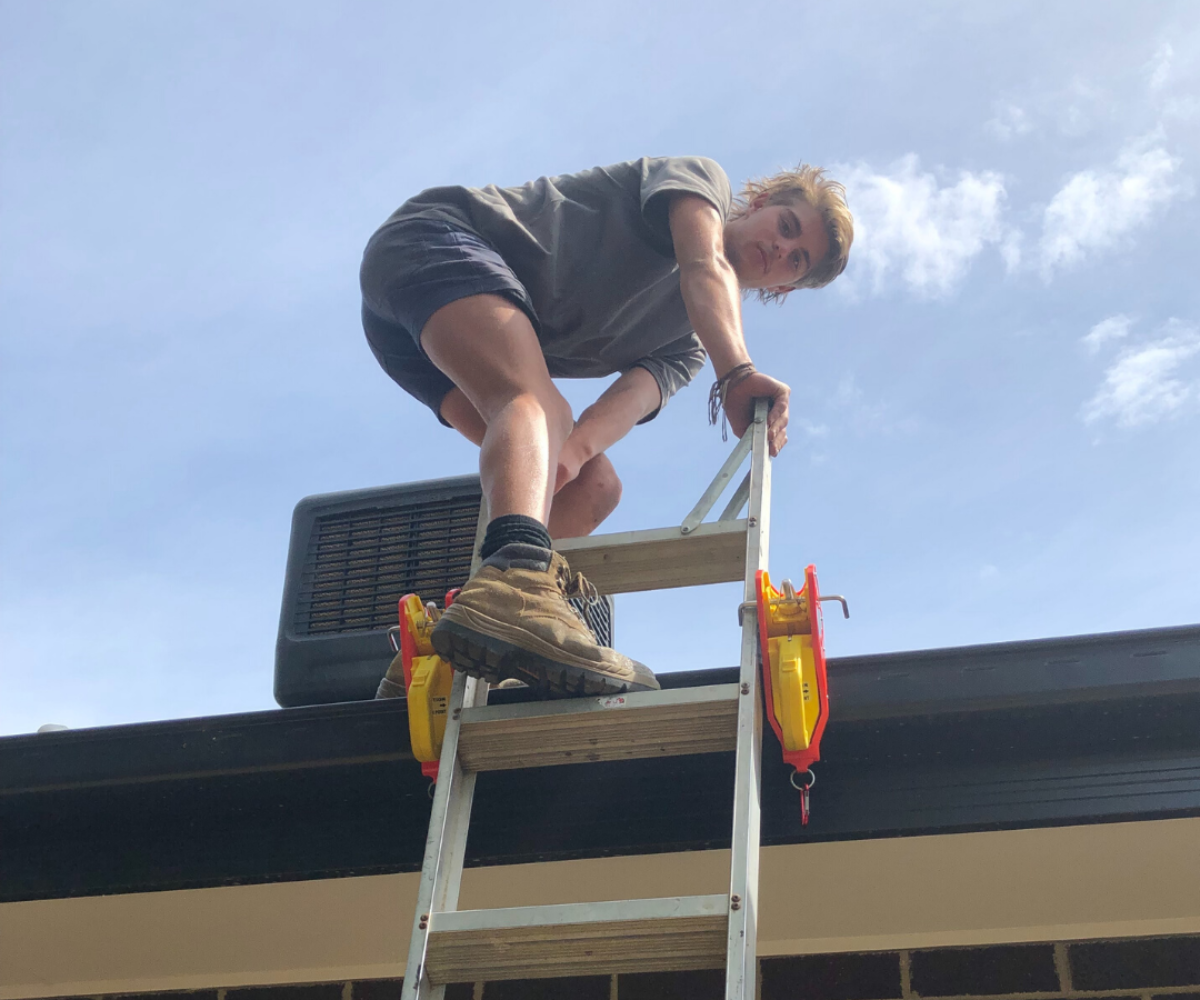  image showcases a worker using a ladder secured with two Lock Jaw Ladder Grip devices, ensuring stability and safety while working on a rooftop.