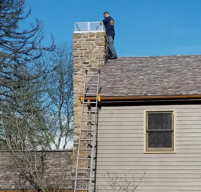 A man is standing on a roof next to a chimney, with a ladder extending from the ground to the roof.
