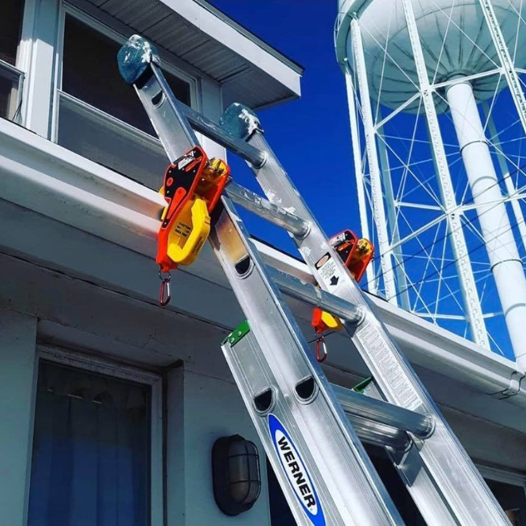A Werner ladder secured to a building's gutter with two Lock Jaw Ladder Grips extends upwards towards a water tower under a blue sky.