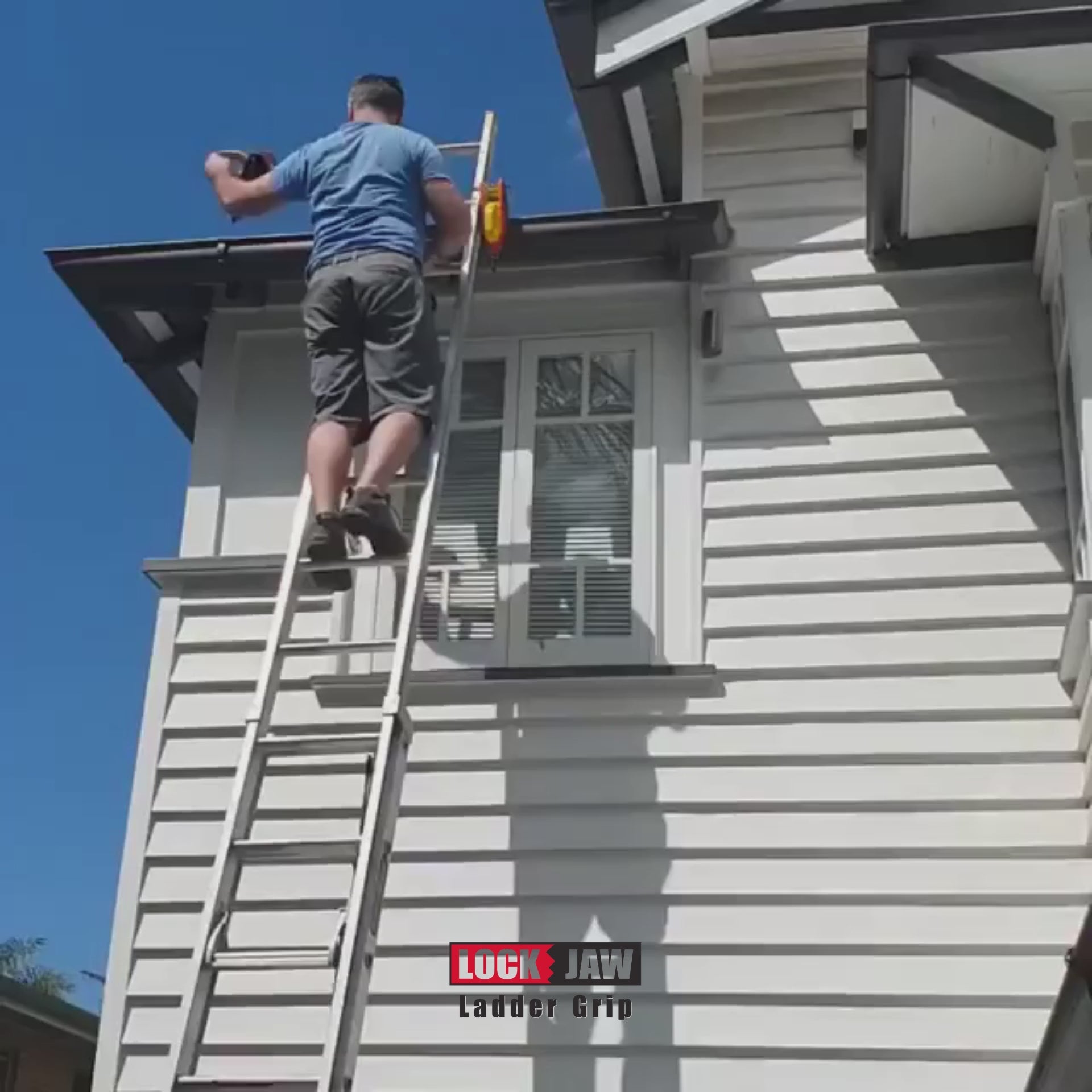A person climbs down a ladder secured to the gutter of a white house with Lock Jaw Ladder Grips under a clear, blue sky.